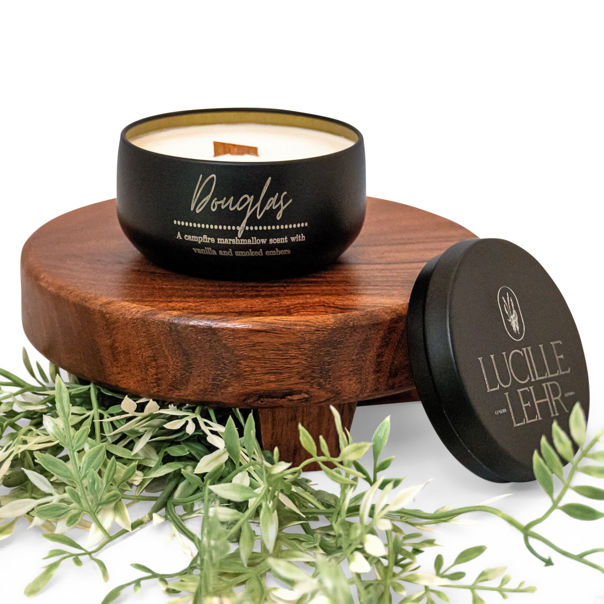 Lucille Lehr's 'Douglas' Candle: Bask in the cozy scent of campfire marshmallows, enhanced with vanilla and smoked embers, with the 'Douglas' candle. Encased in a matte black tin and featuring a crackling wooden wick, this candle rests on a wooden stand adorned with lush greenery, creating a warm and inviting atmosphere in any space.