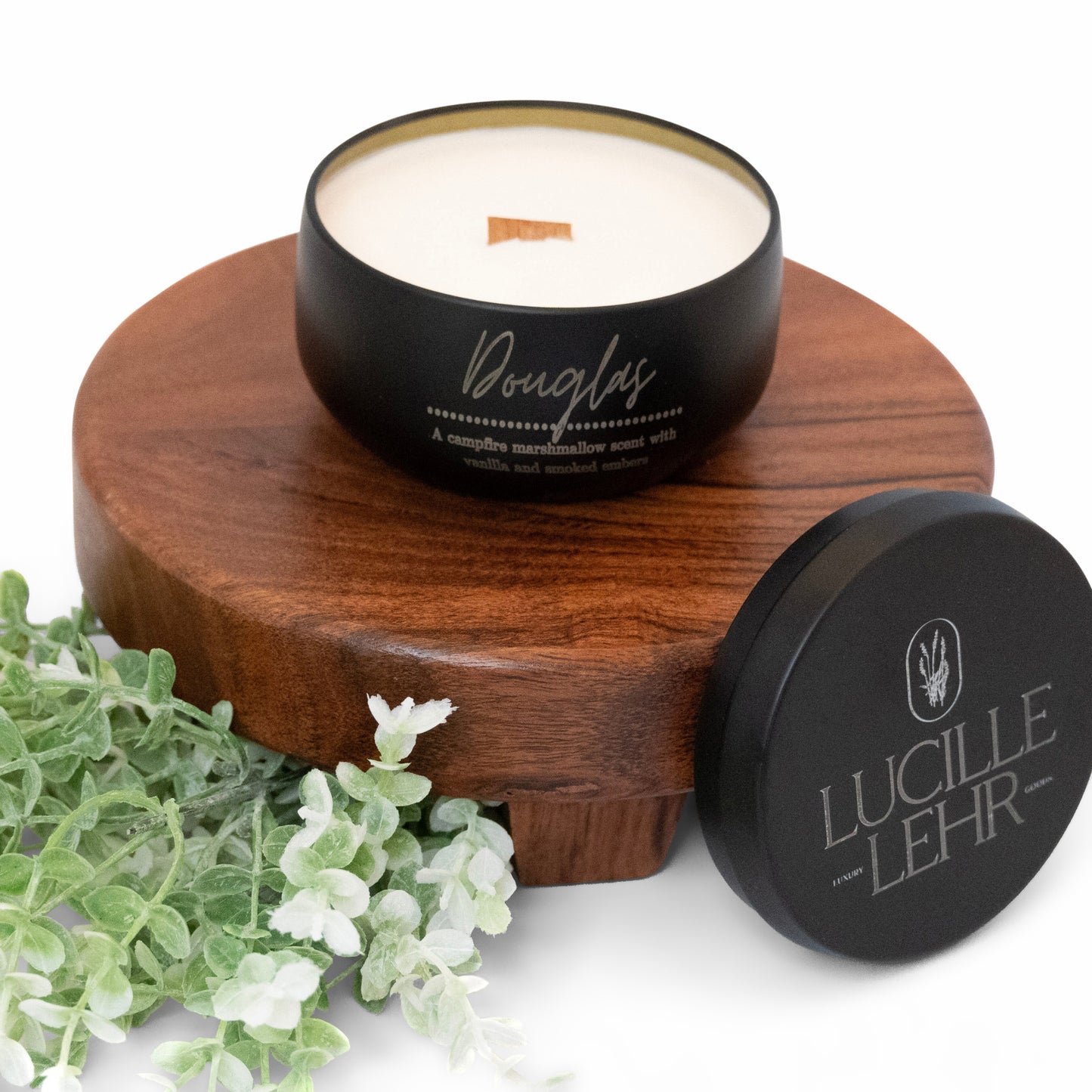 Lucille Lehr's 'Douglas' Candle: Enjoy the comforting aroma of campfire marshmallows with notes of vanilla and smoked embers in the 'Douglas' candle. This matte black tin candle with a crackling wooden wick sits on a rustic wooden stand, complemented by soft greenery, bringing a warm and nostalgic ambiance to any space.