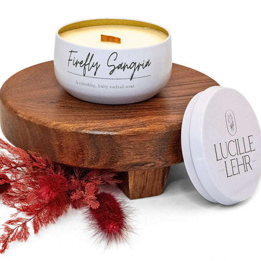 Lucille Lehr's 'Firefly Sangria' Candle: Capture the lively essence of a fruity cocktail with the 'Firefly Sangria' candle, featuring a refreshing, sangria-inspired scent. Presented in a white tin with a crackling wooden wick, it sits on a rustic wooden stand accented by vibrant red dried florals, bringing a playful and invigorating ambiance to any room.