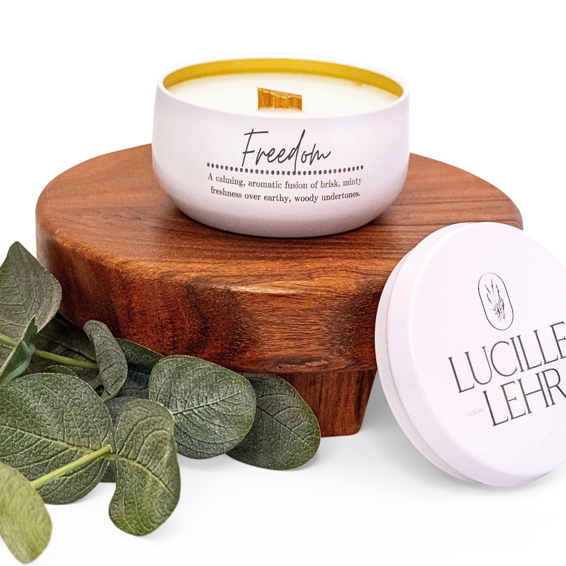 Lucille Lehr's 'Freedom' Candle: Experience a calming blend with the 'Freedom' candle, offering an aromatic fusion of brisk, minty freshness layered over earthy, woody undertones. Housed in an elegant white tin with a wooden wick, this candle sits beautifully on a wooden stand with green eucalyptus leaves, adding a natural, serene touch to your space.