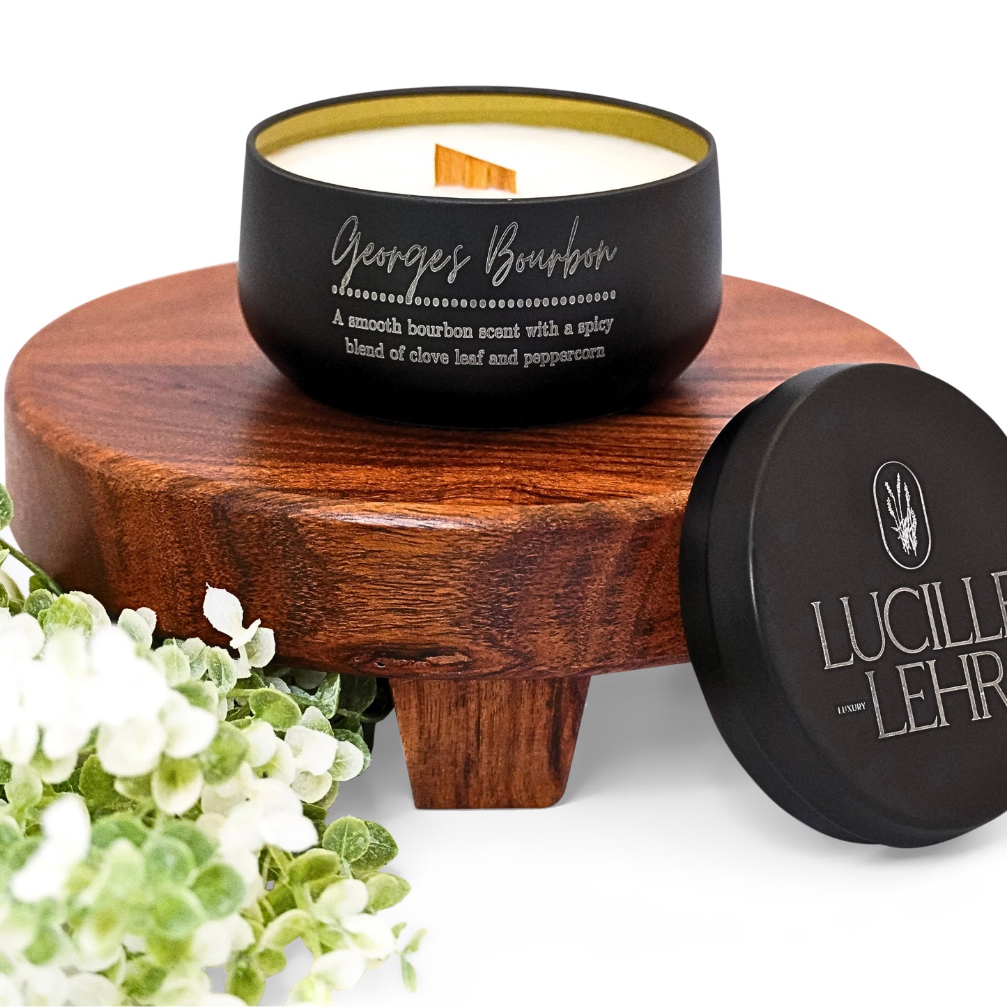 Lucille Lehr's 'Georges Bourbon' Candle: Enjoy the rich, smooth scent of bourbon with spicy hints of clove leaf and peppercorn in the 'Georges Bourbon' candle. Set in a matte black tin with a crackling wooden wick, this candle rests on a rustic wooden stand, accented by soft greenery, bringing warmth and elegance to any setting.