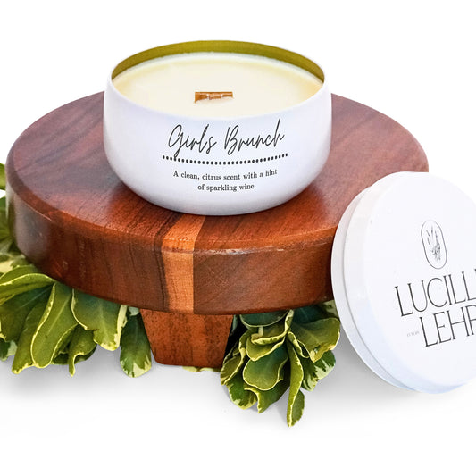 Lucille Lehr's 'Girls Brunch' Candle: Elevate your space with the 'Girls Brunch' candle, featuring a clean, citrus scent with a touch of sparkling wine. Encased in a white tin with a crackling wooden wick, it sits on a wooden stand adorned with fresh greenery, bringing a vibrant and refreshing ambiance to any setting.