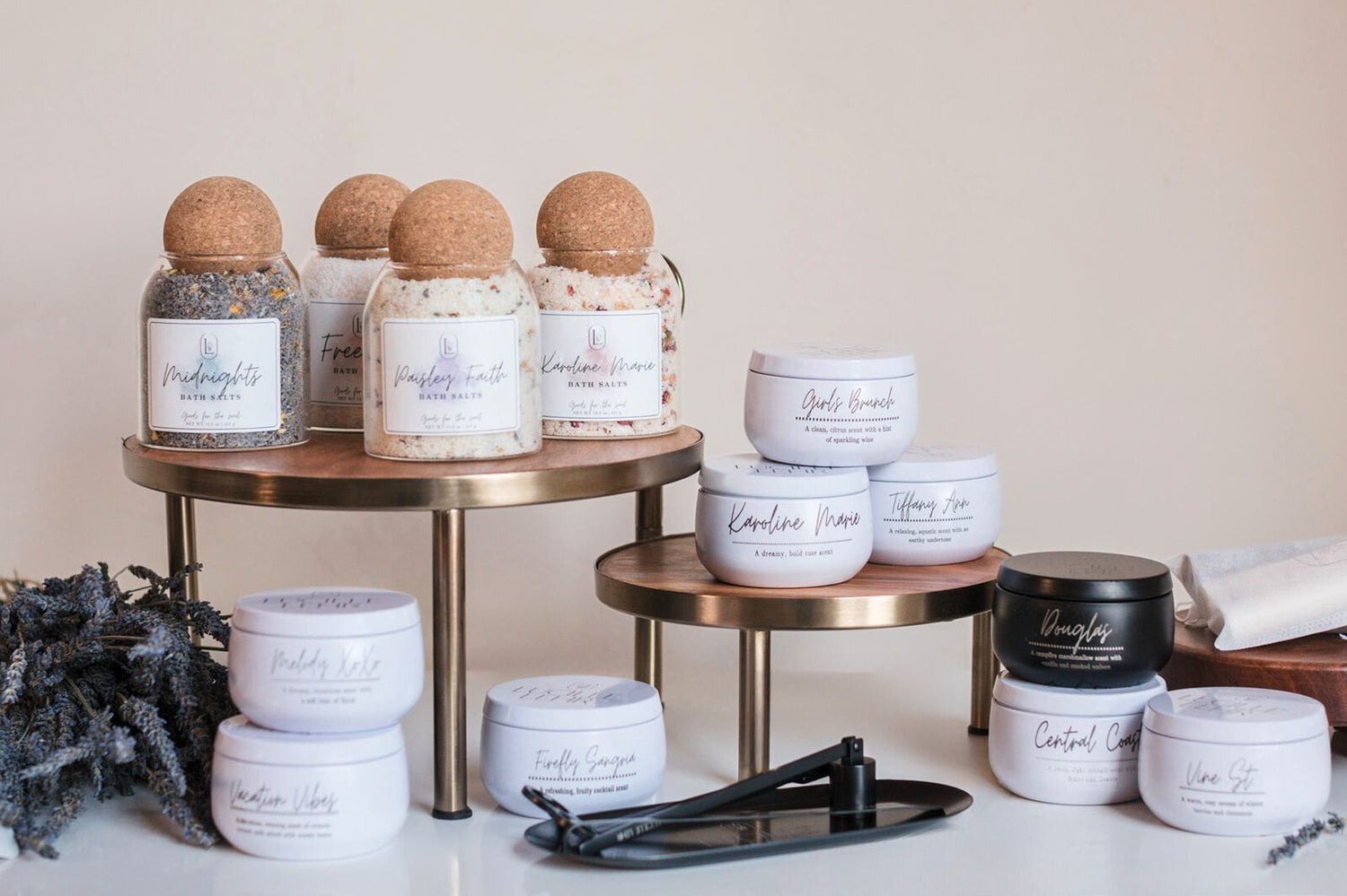 Display of Lucille Lehr Co.'s luxurious lavender-infused products, featuring an assortment of bath salts and candles in elegant white and black tins. Products are arranged on brass stands with a bundle of dried lavender and candle care tools, showcasing a refined and calming aesthetic.