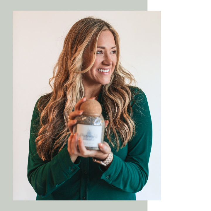 Kandace Slater, maker and owner, smiles while holding a glass jar of bath salts. With her long, wavy blonde hair and wearing a dark green top, the image captures a calm, natural, and wholesome vibe in a minimalist setting.