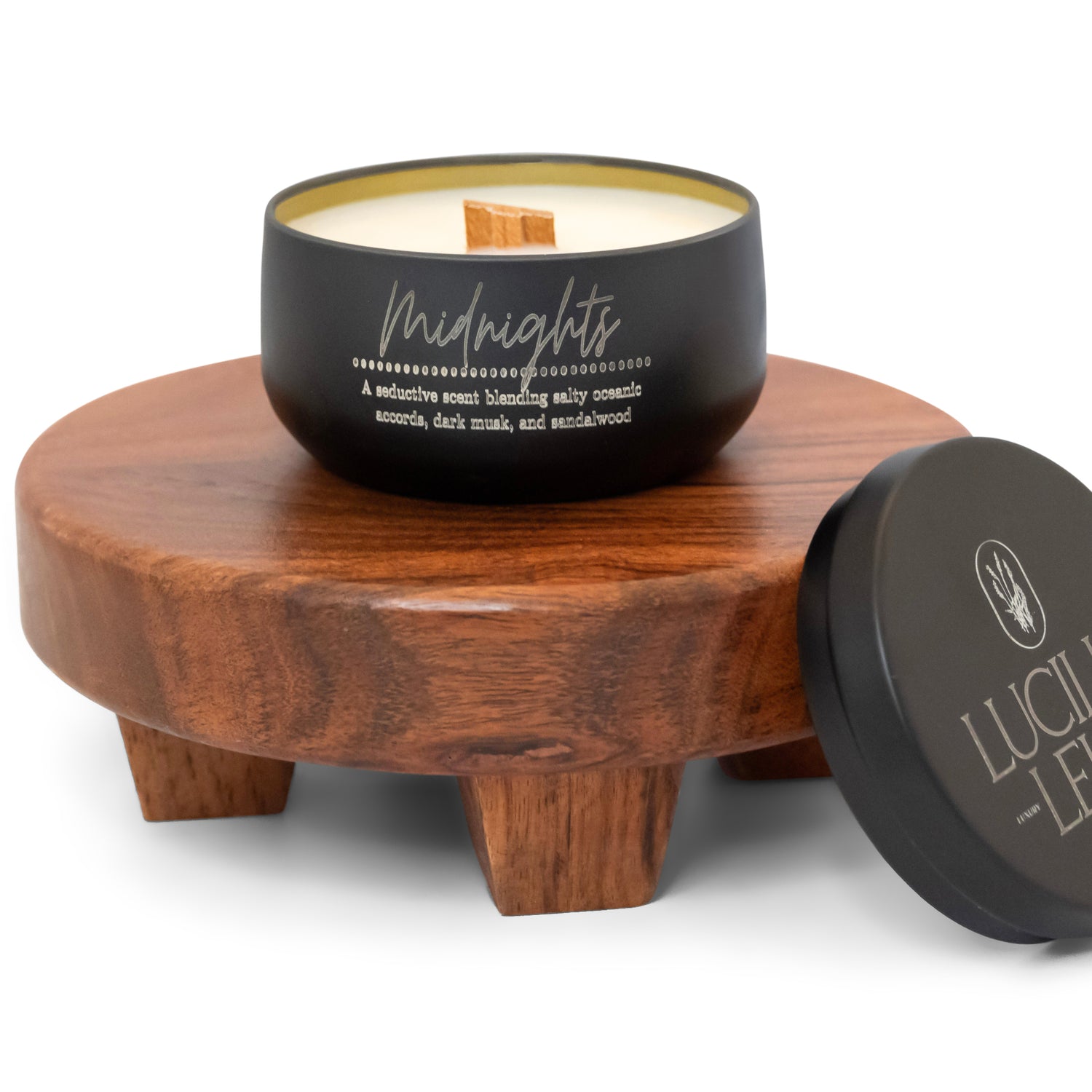 A black candle labeled "Midnights" with a wooden wick sits on a wooden pedestal. Its seductive scent features salty oceanic accords, dark musk, and sandalwood. The lid displays the brand "Lucille Lehr," which has a minimalist, earthy aesthetic.