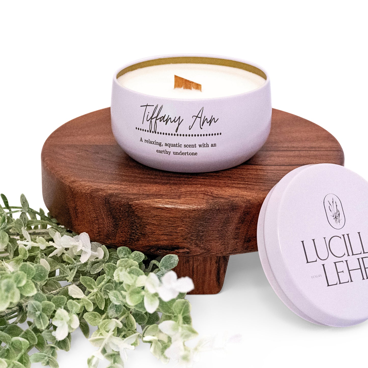 Lucille Lehr's 'Tiffany Ann' Candle: Immerse yourself in a calming, aquatic scent with earthy undertones, hand-poured in a sleek lavender tin featuring a wooden wick for a gentle crackling ambiance. Perfectly placed on a rustic wood stand with delicate greenery, this candle brings an elegant touch to any space.