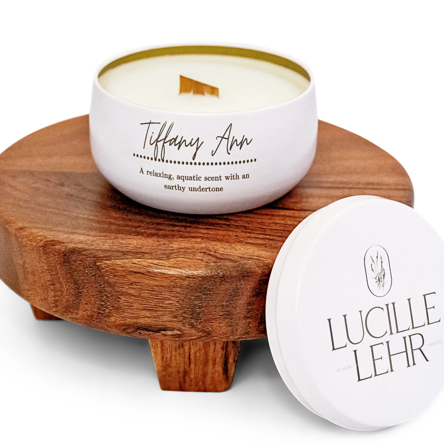 A white candle labeled "Tiffany Ann" with a wooden wick sits on a wooden pedestal. The scent is described as a relaxing, aquatic blend with earthy undertones. The lid beside the candle displays the brand "Lucille Lehr.