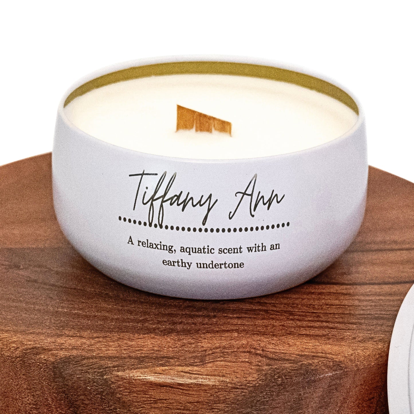 A white candle labeled "Tiffany Ann" with a wooden wick sits on a wooden surface. Its scent is noted as relaxing, aquatic, and earthy.