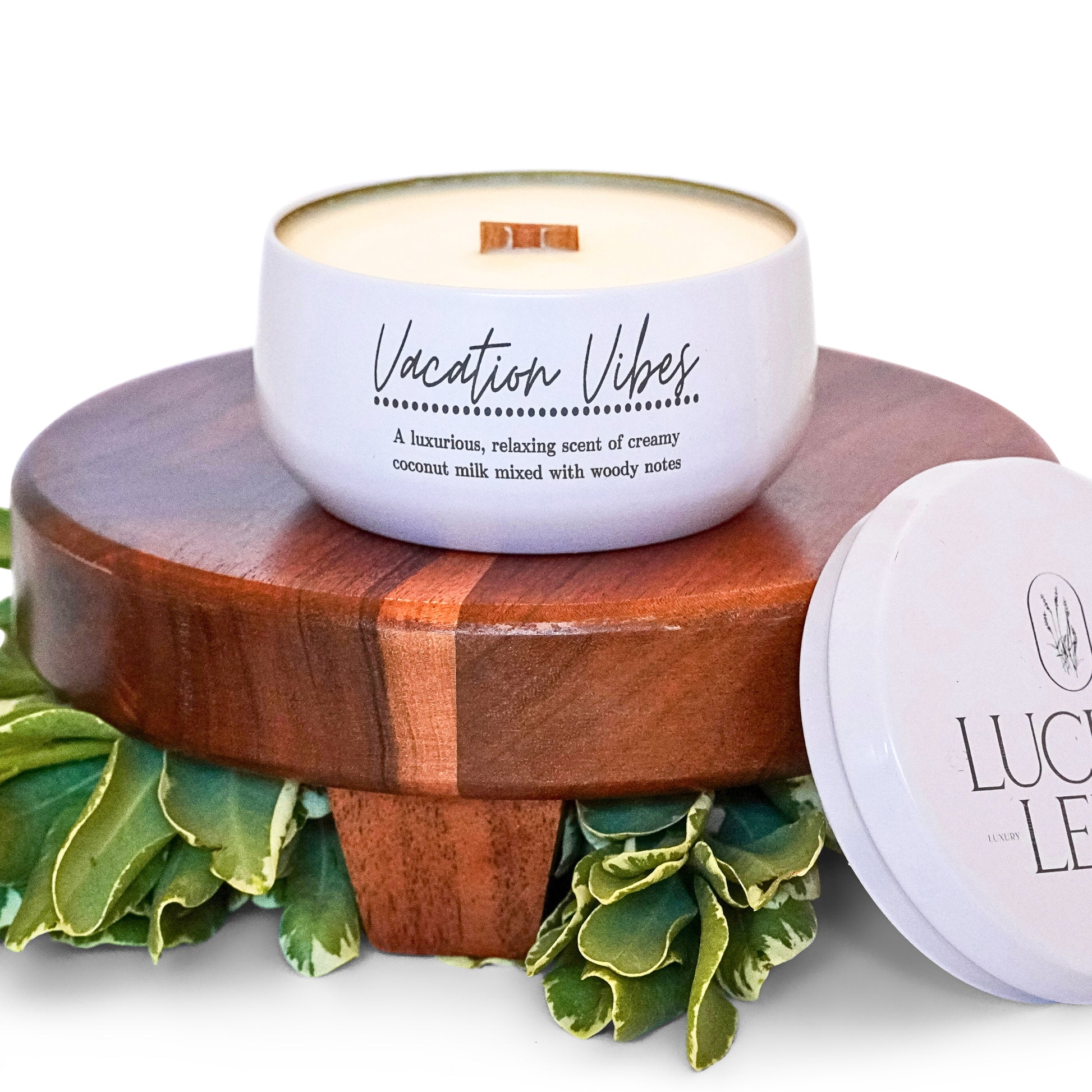 A white candle labeled "Vacation Vibes" with a wooden wick sits on a wooden pedestal, surrounded by greenery. The label describes the scent as a luxurious blend of creamy coconut milk mixed with woody notes. The candle lid, placed beside it, features the brand "Lucille Lehr." The image gives a relaxed and tropical feel, perfect for evoking vacation vibes.