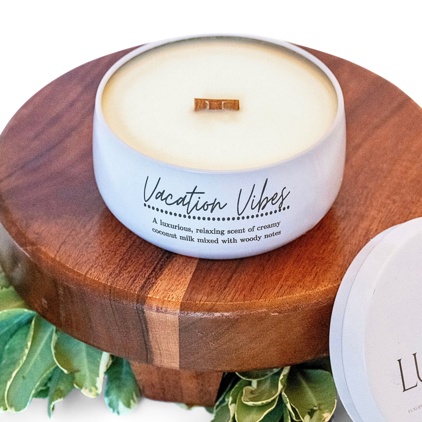 A white candle labeled "Vacation Vibes" with a wooden wick rests on a wooden pedestal surrounded by greenery. The label describes the scent as a luxurious and relaxing blend of creamy coconut milk with woody notes. The overall vibe is calming and tropical, perfect for evoking a vacation atmosphere.