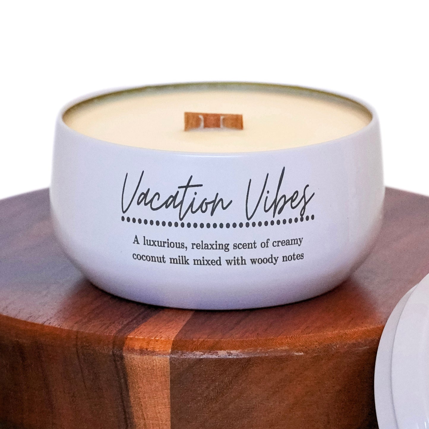 A white candle labeled "Vacation Vibes" with a wooden wick sits on a wooden pedestal. The label describes the scent as a luxurious, relaxing blend of creamy coconut milk mixed with woody notes. The overall aesthetic is clean and calming, evoking a sense of tropical relaxation.